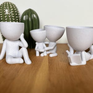 Peut inclure: Quatre pots de fleurs blancs texturés en forme humaine dans diverses poses. L'un est assis, touchant sa tête, un autre s'embrasse et un troisième lit un livre. Deux cactus verts sont en arrière-plan, posés sur une surface en bois.