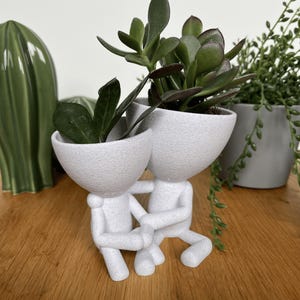 Könnte beinhalten: Ein weißer Keramik-Blumentopf in Form von zwei sich umarmenden Figuren, die sich an den Händen halten. Der Topf ist mit grünen Sukkulenten gefüllt. Weitere Pflanzen in grauen und grünen Töpfen sind im Hintergrund.