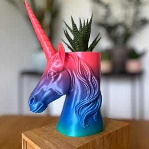 Könnte beinhalten: Ein Einhorn-Kopf-Pflanzgefäß in Rosa-, Lila- und Blautönen, das eine kleine grüne Sukkulente enthält. Der Pflanztopf steht auf einem Holzblock und zeigt das detaillierte Design der Mähne und des Horns des Einhorns.