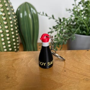 Könnte beinhalten: Ein Miniatur-Schlüsselanhänger in Form einer Sojasoßenflasche. Die schwarze Flasche hat einen weißen Deckel und eine rote Kappe. Die Worte "SOY SA" sind gelb aufgedruckt. Eine silberne Kette und ein Schlüsselring sind befestigt.