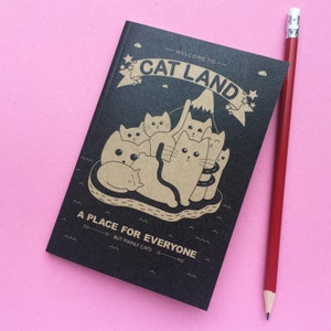 May include: A black notebook with the text "WELCOME TO CAT LAND" and "A PLACE FOR EVERYONE BUT MAINLY CATS" in white and tan. The cover features a cartoon illustration of cats. A red pencil rests beside the notebook on a pink surface.