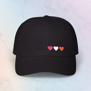 May include: A black baseball cap featuring an embroidered design of three small hearts in a horizontal row. The hearts are colored pink, white, and orange from left to right. The cap has a curved brim and a structured crown with a button on top.