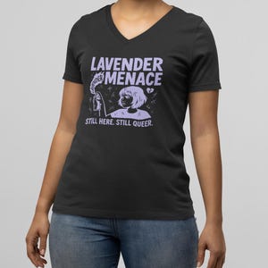 May include: A black V-neck t-shirt featuring a light purple graphic print. The design shows a person holding a spray can with the text LAVENDER MENACE and STILL HERE. STILL QUEER. The graphic includes small symbols like a heart and cross marks.