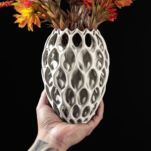 May include: A white ceramic vase with a unique honeycomb design. The vase is being held in a hand and has a bouquet of orange and red flowers.
