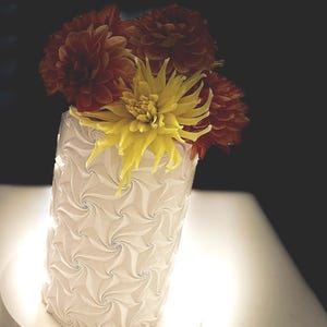 May include: A white vase with a textured, swirling pattern holds a bouquet of dahlias. The flowers are a mix of yellow and reddish-orange, creating a vibrant contrast against the vase. The vase is cylindrical and sits on a white surface.