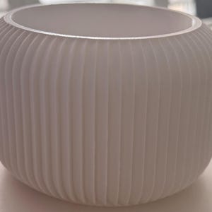 Könnte beinhalten: Eine weiße, zylindrische Vase mit vertikalen, geriffelten Details. Die offene Oberseite zeigt ein glattes Inneres. Die Vase hat einen abgerundeten Boden und ein schlichtes, modernes Design, das sich für die Inneneinrichtung eignet.