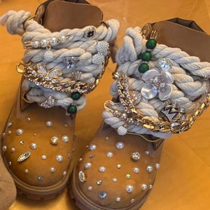 Puede incluir: Un par de botas de cuero color canela con cordones de cuerda blanca gruesa adornados con cadenas doradas, cuentas de perlas, dijes de flores de cristal y imperdibles. Las punteras están decoradas con perlas blancas dispersas y cristales transparentes.
