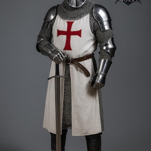May include: A full suit of medieval armor, including a helmet, chainmail, and metal plating. The white tunic features a large red cross. The knight holds a long sword. The armor is worn with black boots and a brown belt.