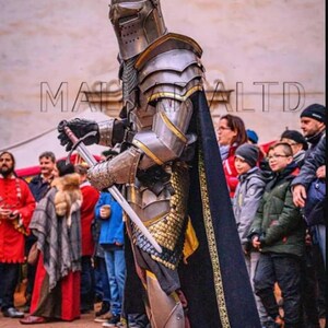 May include: A person in full medieval armor, including a helmet, shoulder guards, and leg protection, holding a sword. The armor is silver and gold, with a black cape. The text "MA... LTD" is visible on the left side of the image.