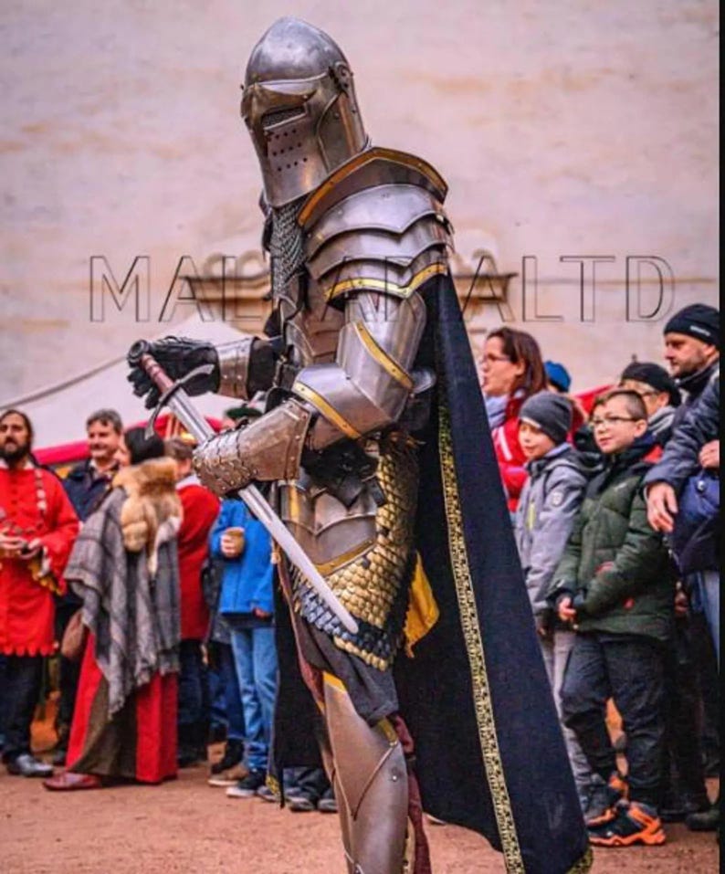 May include: A person in full medieval armor, including a helmet, shoulder guards, and leg protection, holding a sword. The armor is silver and gold, with a black cape. The text "MA... LTD" is visible on the left side of the image.