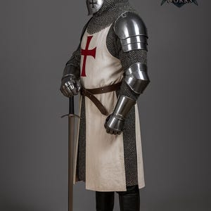 May include: A full suit of medieval armor, including a helmet, chainmail, and metal plating. The white tunic features a large red cross. The knight holds a long sword. The Regal Steel Works logo is visible.