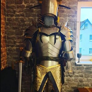May include: A full suit of medieval armor, including a helmet with a chainmail neck guard, shoulder guards, and a chest plate. The armor is silver with gold accents and is displayed against a brick wall. A sword is visible to the left.