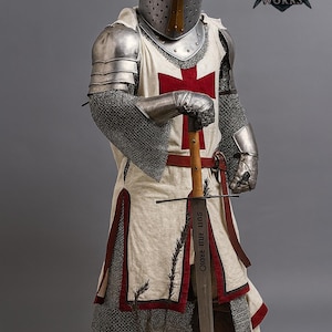 May include: A full suit of medieval armor, including a helmet, chainmail, and a white tunic with a red cross. The knight holds a sword with text on the blade. The armor is silver and gold, and the tunic has red trim.