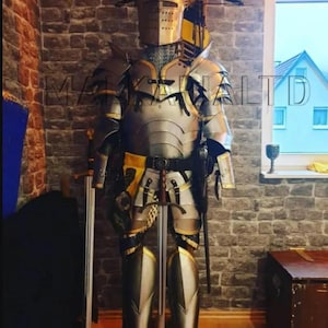 May include: A full suit of medieval armor, including a helmet, chest plate, and leg guards, displayed on a stand. The armor is primarily silver with gold and black accents. Two swords are positioned nearby, suggesting a historical or fantasy theme.