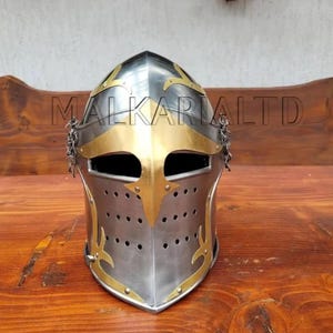 May include: A silver and gold medieval helmet with a riveted construction. The helmet features decorative gold accents, a visor, and a chainmail neck guard. The text "MALKARIALTD" is printed across the top of the helmet.