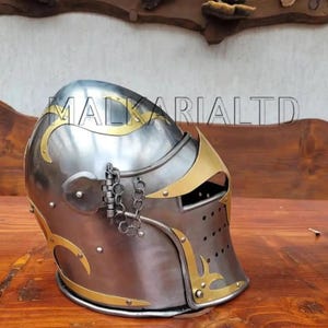 May include: A silver and gold medieval helmet with decorative gold accents. The helmet has a visor with small holes and a hinged section with a chain. The text "MALKARI LTD" is visible on the helmet.