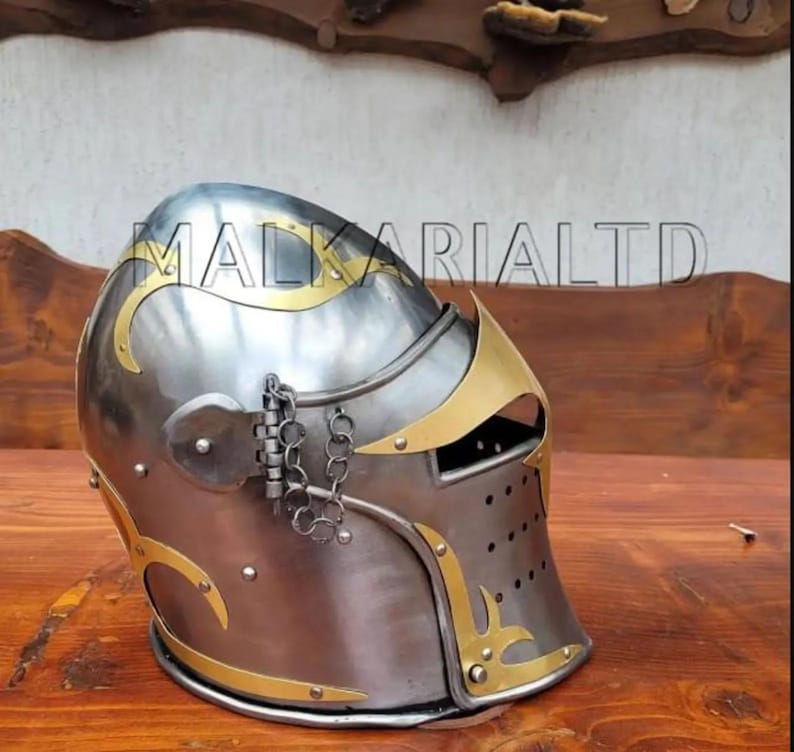 May include: A silver and gold medieval helmet with decorative gold accents. The helmet has a visor with small holes and a hinged section with a chain. The text "MALKARI LTD" is visible on the helmet.