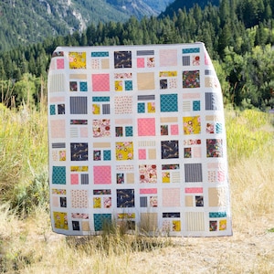 Op de afbeelding: Een wit quilt met een patchwork-ontwerp met vierkanten in verschillende kleuren en patronen, waaronder roze, geel, turquoise en grijs. Het quilt hangt aan een waslijn in een veld.