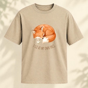 May include: A beige short-sleeved t-shirt featuring a watercolour illustration of a sleeping orange fox curled up next to a small steaming cup. Below the fox, the text "I GO AT MY OWN PACE" is printed in a simple, dark brown sans-serif font.