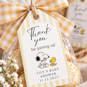 Peut inclure: Une étiquette cadeau rectangulaire à rayures verticales jaunes et blanches, attachée avec un ruban vichy jaune. L'étiquette porte le texte Thank you for joining us!, une illustration de Snoopy et Woodstock avec des sucettes, et LILY'S BABY SHOWER 15.11.2027.