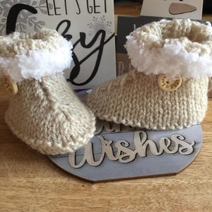 Peut inclure: Une paire de chaussons pour bébé en tricot beige avec une bordure en fourrure blanche et des boutons en bois qui disent "Né en 2023". Les chaussons sont posés sur une pancarte en bois qui dit "Let's Get Cozy" et "Warm Wishes".
