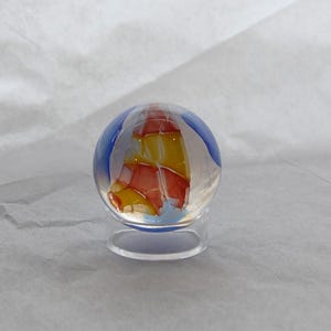 May include: A clear glass marble with a colorful design. The marble features a blue, yellow, and red pattern, resembling a sailboat. It sits on a clear, circular stand, showcasing the intricate artwork. The marble is a decorative item.