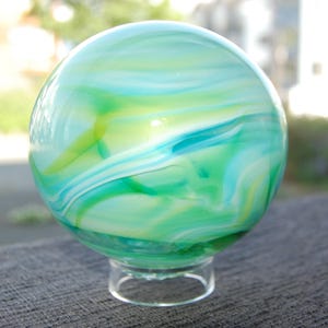 May include: A decorative glass sphere with a clear glass base. The sphere is swirled with shades of green, blue, and white. The glass has a glossy finish and appears to be hand-blown. The sphere is a decorative item.