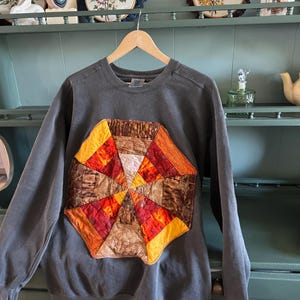 May include: A charcoal gray crewneck sweatshirt featuring a large, central, octagonal quilt-style appliqué. The appliqué is composed of triangular fabric segments in shades of orange, yellow, brown, and deep red, arranged in a radial pattern.