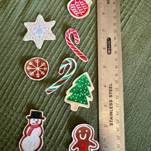 Puede incluir: Un conjunto de siete parches de fieltro con motivos navideños que incluyen un adorno rojo, un copo de nieve, dos bastones de caramelo, un árbol verde, un muñeco de nieve y una galleta de jengibre, junto a una regla de acero de 30 cm.