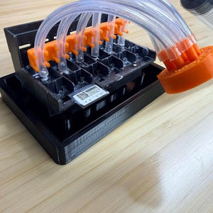 Puede incluir: Un dispositivo de plástico negro con conectores naranjas y tubos transparentes, conectado a una jeringa. El dispositivo se encuentra sobre una base negra, con una pequeña etiqueta visible. La configuración está sobre una superficie de madera clara.