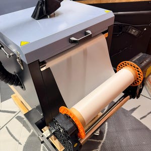 Op de afbeelding: Een industriële sublimatieprinter met een lichtblauw bovenstuk en zwarte accenten. Een rol wit transferpapier is aan de voorkant gemonteerd. De machine heeft een oranje en zwart rollersysteem en een zwarte uitlaatslang.