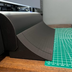 Op de afbeelding: Een zwarte plastic helling met een gebogen rand, die op een groene snijmat en een houten oppervlak ligt.