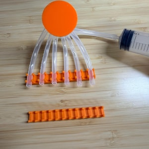 Op de afbeelding: Een oranje kunststof verdeelstuk met zes doorzichtige buizen verbonden met een oranje schijf. Een spuit is aangesloten op een van de buizen. Een extra oranje kunststof stuk ligt eronder. Op een houten oppervlak.