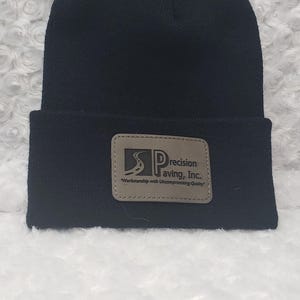 May include: Black knit beanie with a folded brim. A rectangular patch on the front displays the text "Precision Paving, Inc." and the tagline "Workmanship with Uncompromising Quality."