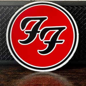 May include: A circular sticker featuring the Foo Fighters logo. The design includes the letters "FF" in black, set against a vibrant red background. The logo is outlined with a thin white border.