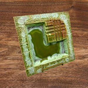 Puede incluir: Un cenicero de cerámica verde y dorado con un diseño geométrico. El cenicero tiene forma cuadrada y un borde elevado.