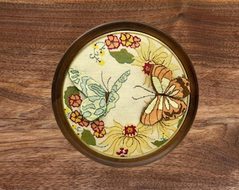 May include: A round embroidery hoop with a white background and a floral design featuring two butterflies. The butterflies are embroidered in shades of blue, green, orange, and yellow. The flowers are embroidered in shades of red, yellow, and green.