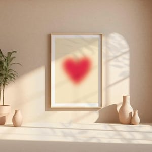 May include: A framed print of a blurred red heart on a beige background, with a potted plant and two ceramic vases on a white surface.