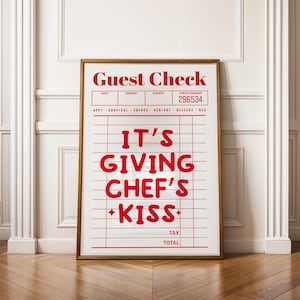 May include: A framed print with a white background and red text that reads "Guest Check" and "IT'S GIVING CHEF'S KISS". The print is in a gold frame and has a grid pattern.