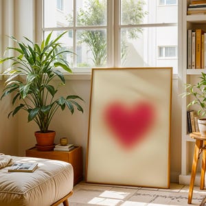 May include: A framed print of a blurred pink heart shape on a white background. The print is hanging on a wall in a room with a plant and a window.