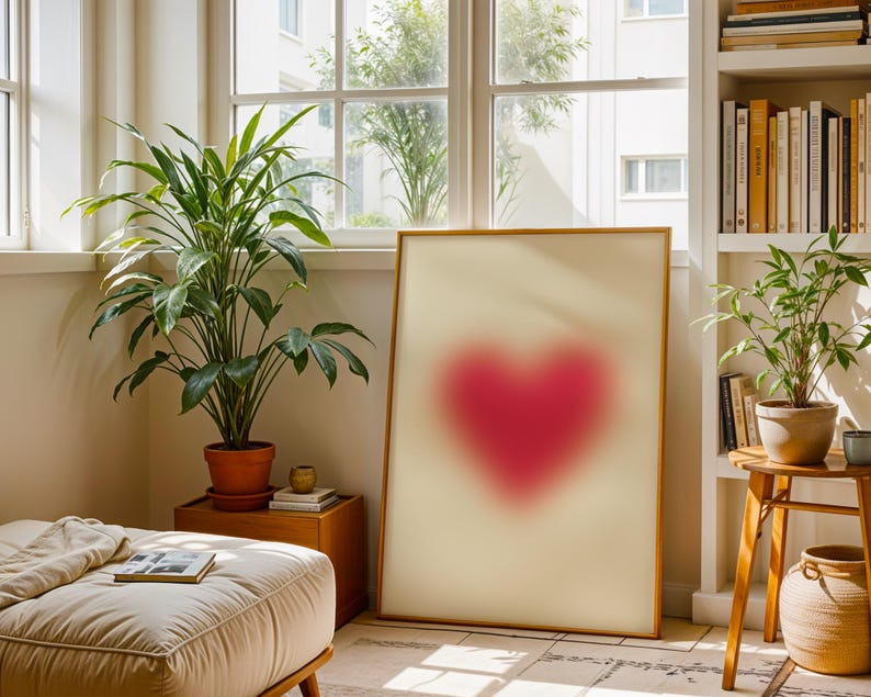 May include: A framed print of a blurred pink heart shape on a white background. The print is hanging on a wall in a room with a plant and a window.