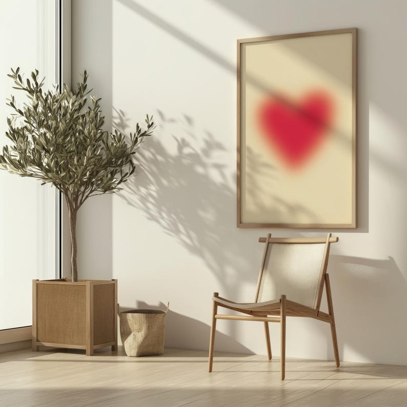 May include: A framed print of a red heart shape on a beige background, hanging on a white wall. A wooden chair with a white cushion sits in front of the print. A potted olive tree is in the corner of the room.
