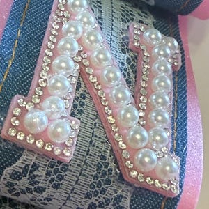 May include: A decorative letter N patch featuring a light pink felt base adorned with white faux pearls and clear rhinestones. The letter is layered over a strip of blue denim fabric with gold stitching and a white floral lace overlay trim.