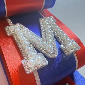 May include: A decorative ribbon accessory featuring a large letter M and a partial letter T, both embellished with white faux pearls and clear rhinestones. The base consists of layered royal blue and bright red satin ribbons with silver glitter trim.