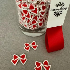 May include: A glass filled with red heart-shaped embellishments trimmed with white pearls and a central rhinestone. Three similar embellishments are scattered nearby. A red satin ribbon and a tag with the text "Mum-a-licious" are also present.