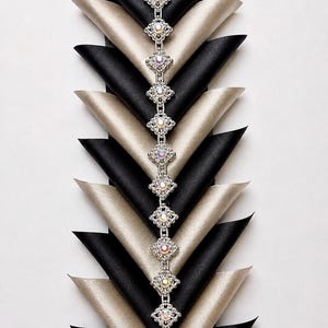 May include: A vertical decorative trim featuring a central chain of silver-toned, diamond-shaped links, each set with a small iridescent crystal. This chain is flanked by alternating, folded fabric triangles in solid black and champagne beige.