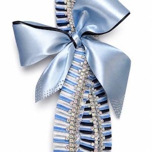 May include: A decorative homecoming mum accessory featuring a large light blue satin bow with black edges. The vertical streamers consist of pleated ribbons in light blue, white, black, and silver glitter, accented by vertical rows of clear rhinestones.