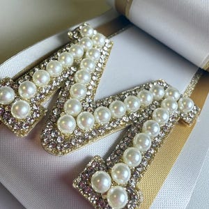 May include: A decorative letter M applique embellished with white faux pearls and clear rhinestones, mounted on a layered white and gold ribbon base. The letter features a gold-toned border and is centered on the smooth, folded fabric ribbons.
