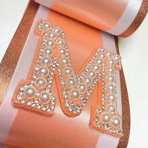 May include: A hair bow featuring a large letter M embellished with white faux pearls and clear rhinestones. The bow is constructed from layered ribbons in shades of peach, white, and metallic copper, creating a multi-textured appearance.