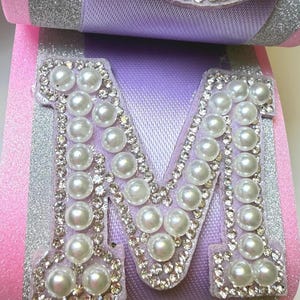May include: A decorative letter M crafted from lavender ribbon with silver glitter accents. The letter is embellished with a border of small clear rhinestones and filled with white faux pearls. A small rectangular piece below also features pearls.